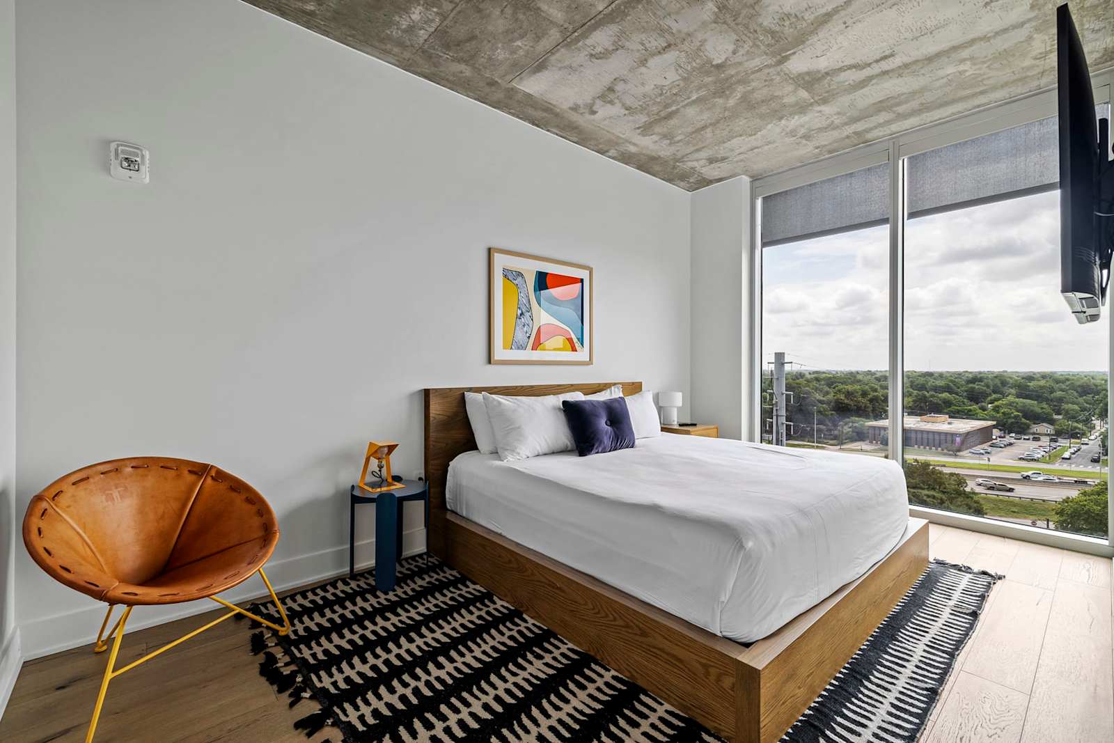 Queen bed in unit 1204 beside floor-to-ceiling windows, with a rattan lounge chair and a wall-mounted TV