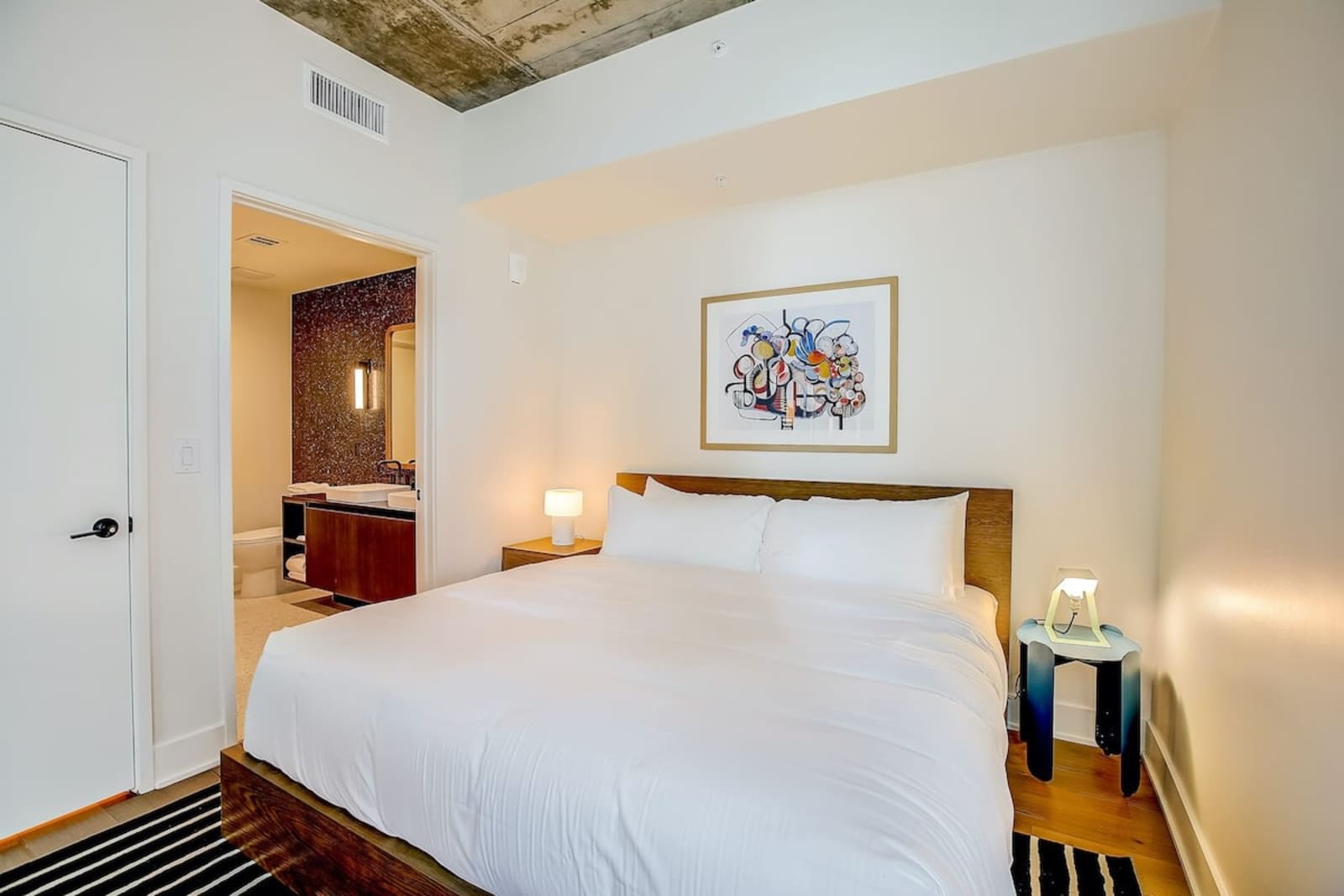 Second bedroom in unit 1406 with a queen bed, bedside lamp and a doorway through to the bathroom