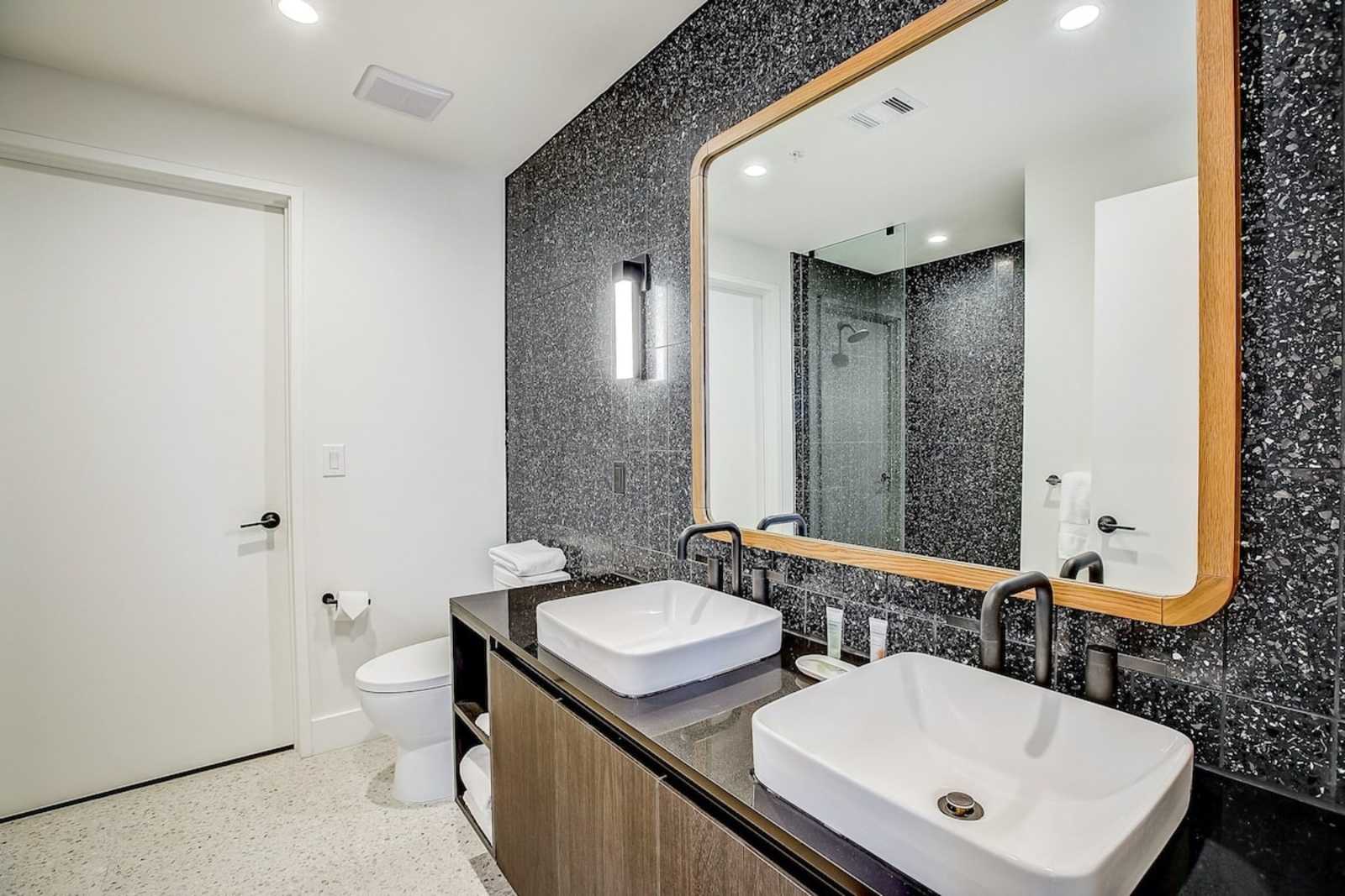 Bathroom in unit 1406 with twin vessel sinks, a brass-framed mirror and dark textured tile