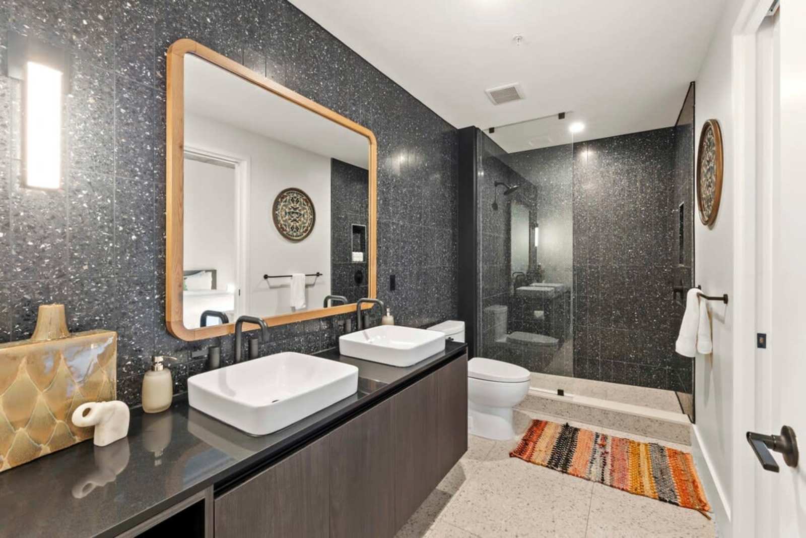 Bathroom in unit 1804 with twin vessel sinks, a brass-framed mirror, walk-in shower and patterned rug